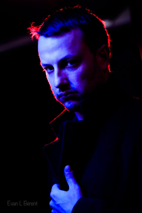 A portrait of a stern-faced man light by blue lights, while backlit by red light.
