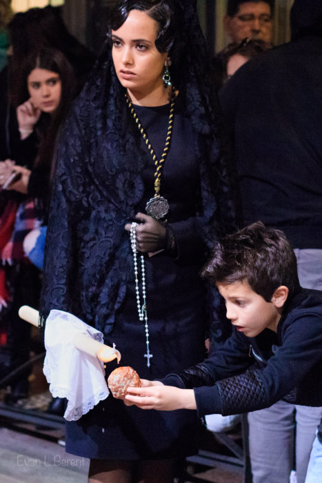 A woman wearing a mantilla looks to the left while a boy collects the dripping wax.